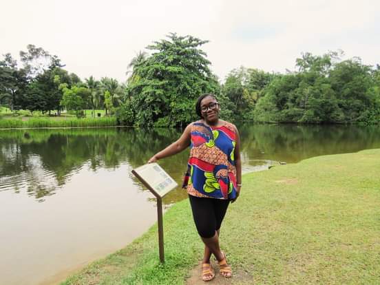 Anike standing by a peaceful lake with information plaque in a green park setting