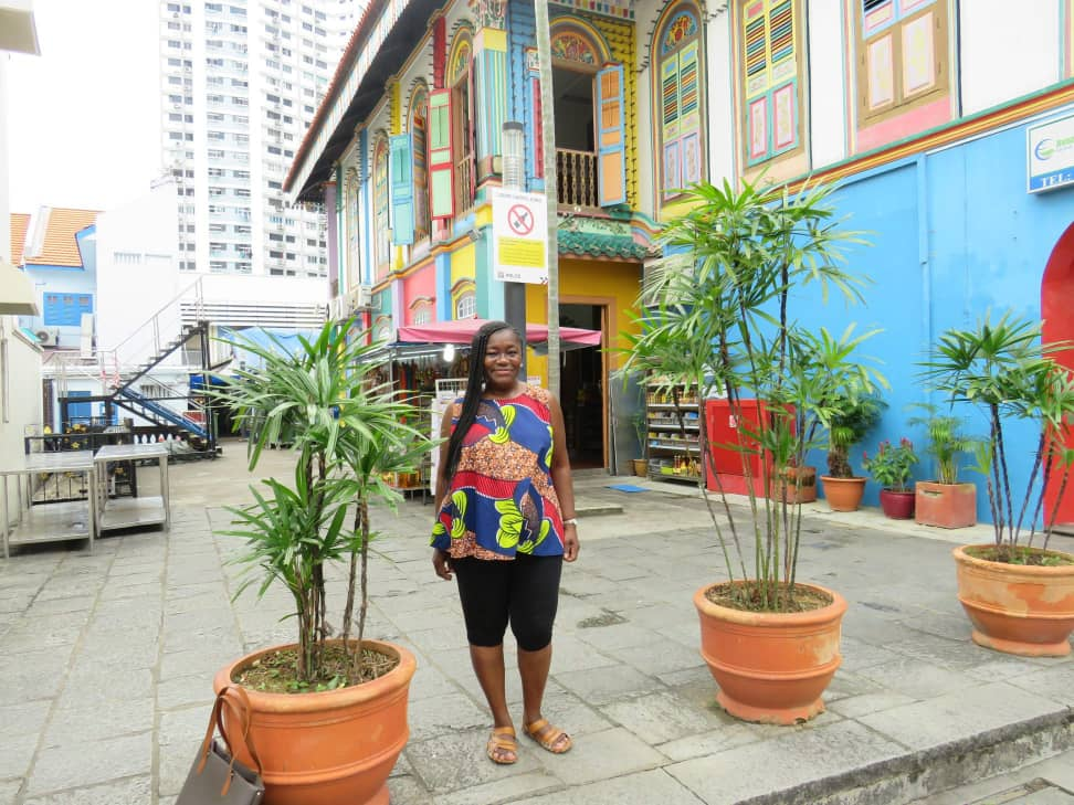 Anike standing in front of a colorful traditional shophouse with vibrant decorations and potted plants