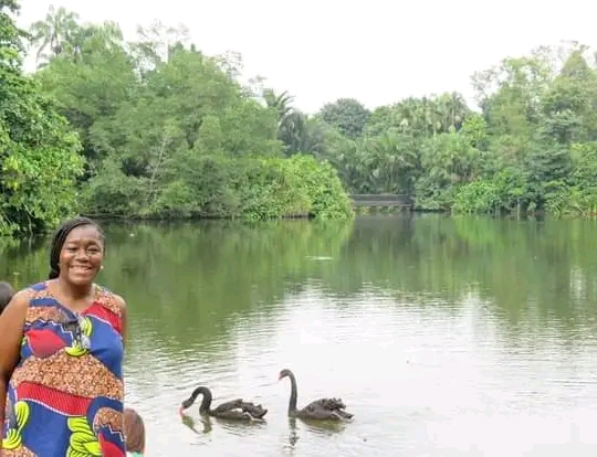 Anike standing by a serene lake with black swans swimming in the water surrounded by lush greenery